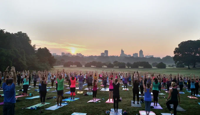 Large group practicing yoga outdoors at sunrise with city skyline in the background.