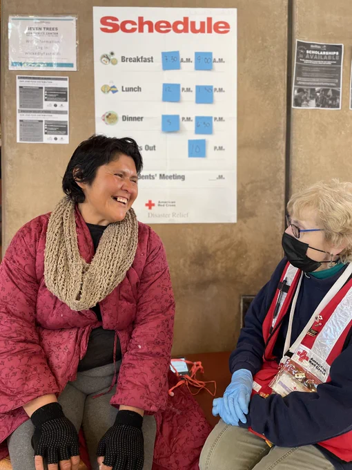 Two people are sitting and smiling at each other in front of a wall with a schedule. One is wearing a red jacket with an American Red Cross logo, gloves, and a face mask. The other person is wearing a red coat and beige scarf. The background shows a schedule for meals and a Red Cross Disaster Relief poster.