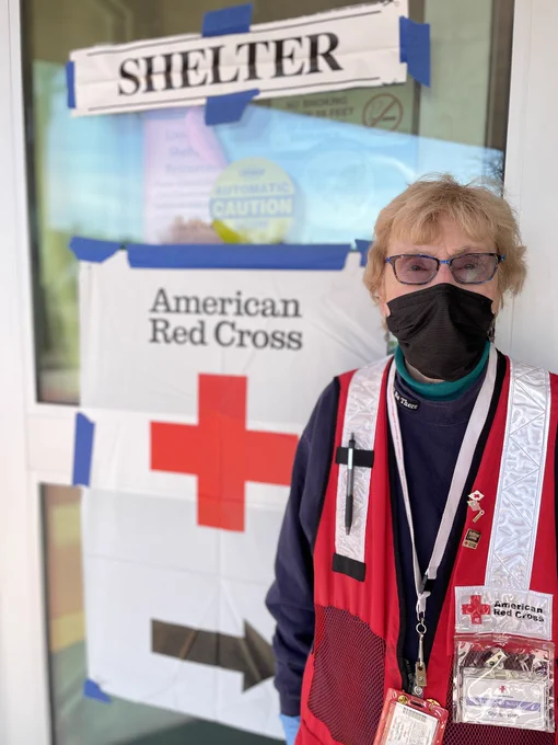 A person with a mask stands in front of a glass door with a sign labeled "Shelter" above and an American Red Cross banner with a large red cross on it. They are wearing a red vest with Red Cross insignia and multiple lanyards.
