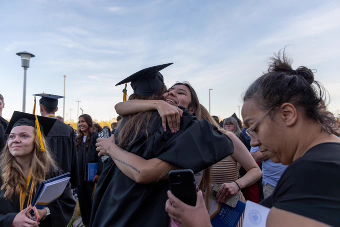 Graduates in caps and gowns celebrate with hugs and smiles outdoors. Onlookers include family members taking photos. The scene is lively and joyful.