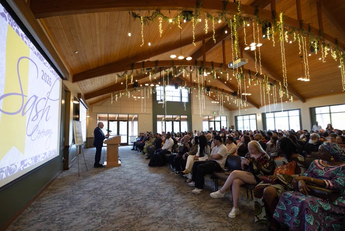 A speaker stands at a podium addressing a large audience seated in a spacious venue with wooden ceilings and hanging greenery. A large screen displays "2026 Sash Ceremony." The room is well-lit, with many attendees listening attentively.