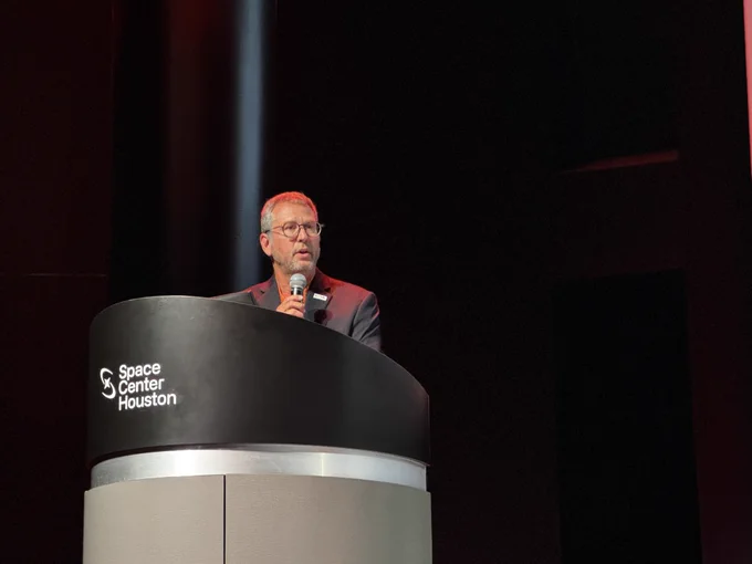 A person is speaking at a podium labeled "Space Center Houston," holding a microphone and standing against a dark background.
