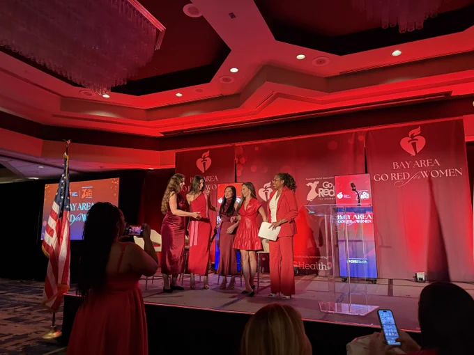 A group of people dressed in red are standing on a stage under bright red lighting. They are in front of banners that read "Bay Area Go Red for Women." An American flag is displayed to the left.