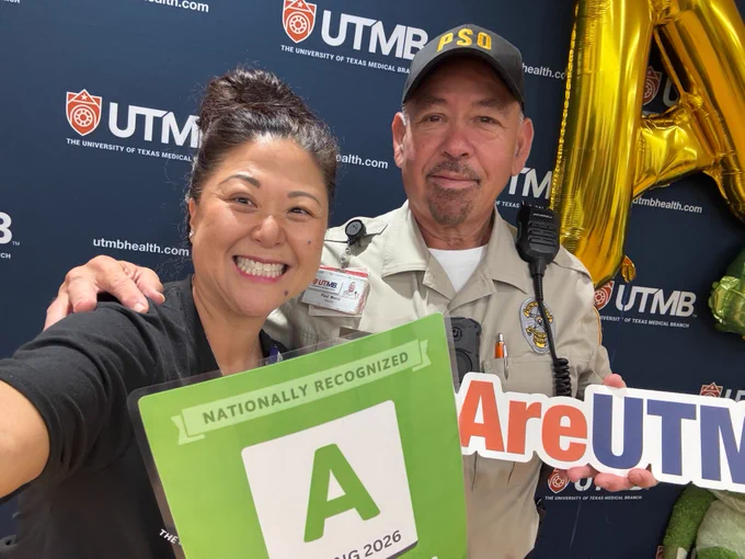 Two individuals are smiling in front of a backdrop with the University of Texas Medical Branch (UTMB) logo. One is holding a green sign with an "A" rating for Spring 2026, and the other is holding a sign saying "We Are UTMB." There are golden balloons partially visible in the background.