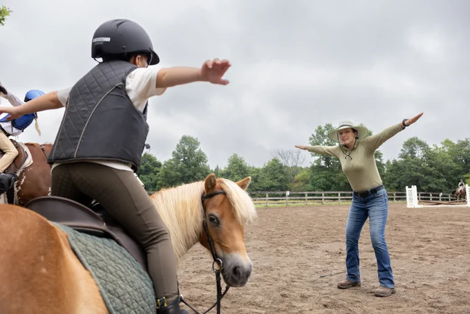 A person on horseback stretches out their arms, mirroring a person standing in front who is also extending their arms. The scene takes place in a sandy riding arena with trees and a fence in the background.