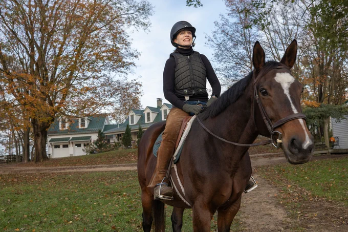 A person is horseback riding on a brown horse in front of a large house with a green roof. The scene is outdoors, surrounded by trees with autumn foliage.