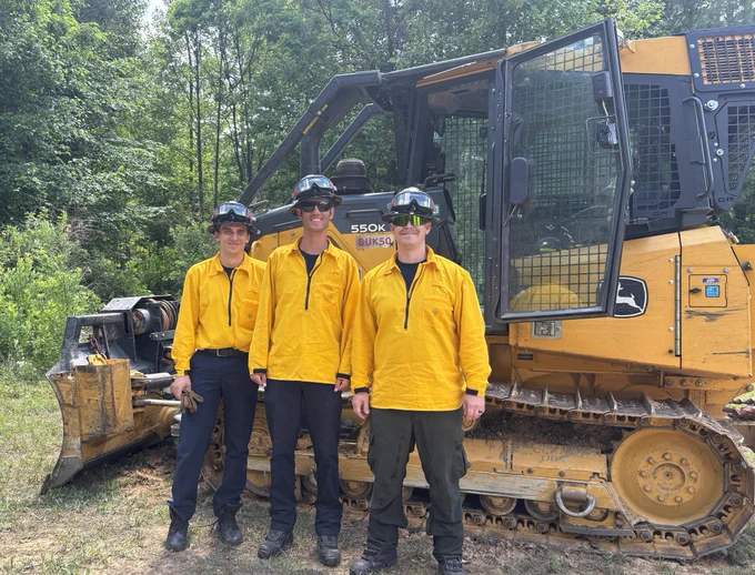 Three people in yellow uniforms stand in front of a John Deere 550K bulldozer in a forested area. They are wearing helmets with visors.
