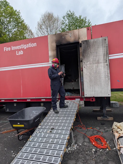 A person in protective gear and a helmet stands on a metal ramp leading to the open door of a red trailer labeled "Fire Investigation Lab." The trailer shows some signs of fire damage. A wheelbarrow and electrical cords are visible nearby. Trees are in the background.
