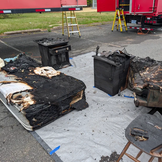 A collection of charred furniture pieces, including a chair and small tables, sits on a tarp in a parking area. In the background, red trucks and ladders can be seen.