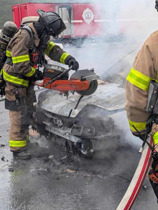 Firefighters are using a power saw to cut open the hood of a vehicle with visible smoke. A firetruck is parked in the background.