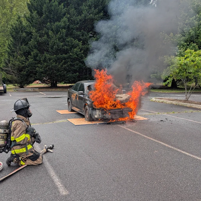 A firefighter, wearing protective gear, kneels on the ground and sprays water to extinguish flames engulfing the front of a burning car. Thick black smoke rises from the fire. The scene takes place in a parking lot surrounded by lush green trees.
