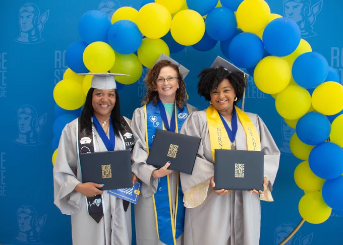 Three RSC graduates take a photo in front of a yellow and blue balloon arch. They are wearing caps and gowns while holding diplomas.
