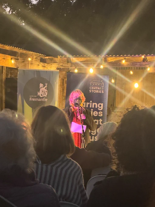 Picture from further away with lights and crowd in foreground - Nicola is a white woman, wearing red dress, black cardigan, emu feather earrings and a black KN95 mask decorated with temp tattoo leaf design. She’s speaking into a mike with a sign behind her that’s says centre for stories