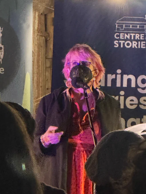 Nicola is a white woman, wearing red dress, black cardigan, emu feather earrings and a black KN95 mask decorated with temp tattoo leaf design. She’s speaking into a mike with a sign behind her that’s says centre for stories