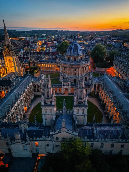 Aerial view of Oxford at blue hour, centred on the Radcliffe Camera and All Souls College in the foreground. Warm lights glow across the historic city as the sun sets on the horizon, with Oxford’s spires and rooftops stretching into the distance.