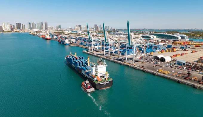 Aerial view of the port. A container ship named \"WARNOW WHALE\" is being assisted by a tugboat. The docks are filled with shipping containers and large cranes. The city skyline is visible in the background.