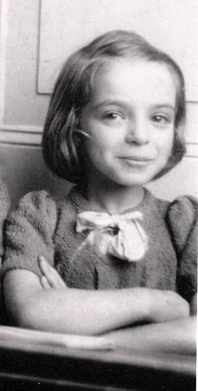 A vintage black and white photo of a young girl smiling, wearing a sweater with a bow on the collar, arms crossed in front of her.