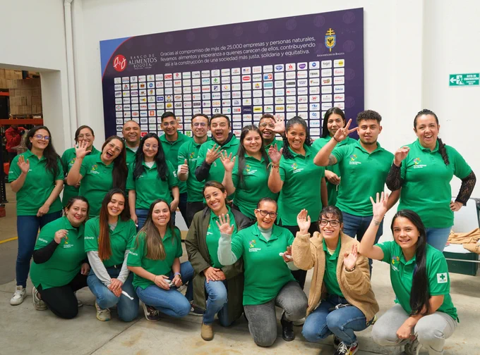 Grupo de personas sonrientes con camisetas verdes posando y saludando frente a un cartel lleno de logotipos en la pared. Están en un ambiente laboral, transmitiendo un sentimiento de camaradería y entusiasmo.