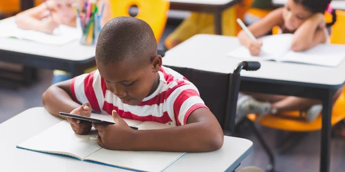 A young student wearing a red and white striped shirt sits at a desk while looking at a tablet screen.