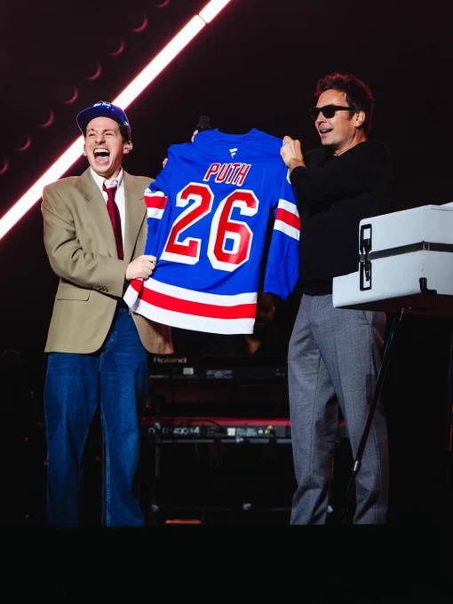 Charlie Puth and Jimmy Fallon on stage. Both are holding up a Rangers jersey. 
