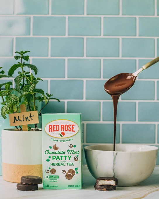 Box of Red Rose Chocolate Mint Patty Herbal Tea next to fresh mint plant and chocolate-covered mint patties on a white countertop.