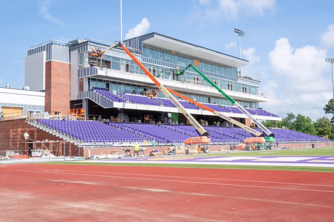 A photo of Tennessee Tech's Tucker Stadium under construction with a large, modern grandstand featuring purple seating. Two construction lifts are positioned in front, with workers visible. The stadium has a running track and clear blue sky overhead.