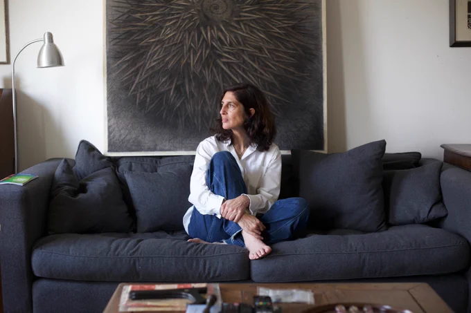 Catherine Kurtz sits on a sofa with her legs crossed. She wears a white shirt and blue jeans. She looks out of a window, out of frame.