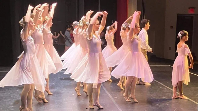 A group of ballet dancers in pink dresses and shoes perform on stage. The dancers have their arms raised above their heads and they are all facing the same direction.