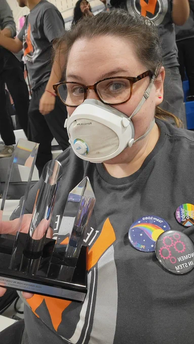 OP holding a trophy from the FIRST Robotics Competition, in her team uniform and wearing a Flo Mask with a sip valve. She has three buttons pinned to her T-shirt: one reads "women in STEM," one is a competition-themed pride button, and one is a team-specific pride button.