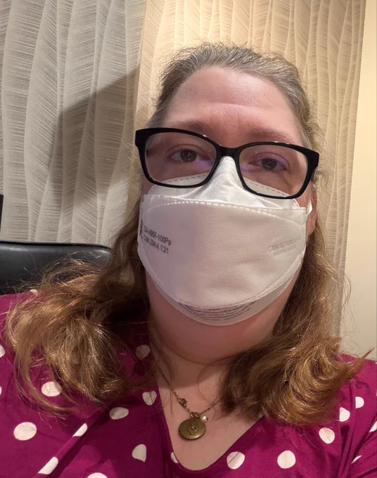 Woman in a purple top with white polkadots, mid-length brown hair, a gold pendant necklace and glasses is wearing a Canadamasq Q100 respirator (N95).