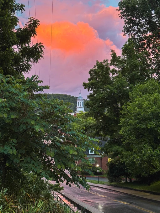 A scenic view of Jefferson Hall surrounded by lush green trees. The sky features vibrant orange and pink clouds during sunset, adding a warm glow to the scene. A road runs through the foreground, slightly wet from recent rain.