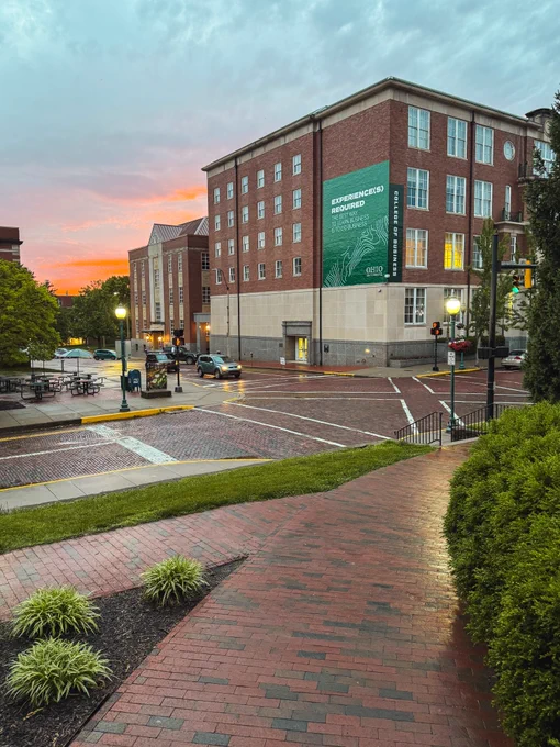 A city intersection at sunset with brick buildings and streets, and a large Ohio University sign on the building. Street lamps are lit, and there are parked cars and trees lining the sidewalks.