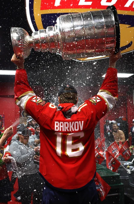 barkov lifting the stanley cup in the panthers locker room while being sprayed with champagne