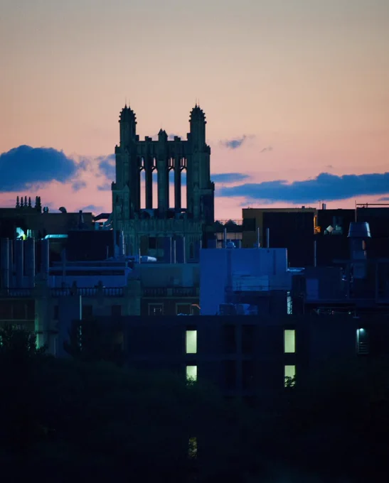 Silhouetted gothic tower rising above city buildings at dusk with a pink and blue sky.