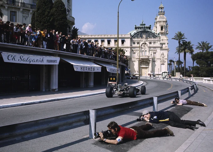 Shot from the outside of Massenet corner, on the bottom right of frame, three photographers lie on their fronts, taking photographs of cars coming up the hill from under the crash barrier. Across the track, spectators line the balcony above a row of shops, watching cars pass. 1968 Monaco Grand Prix.