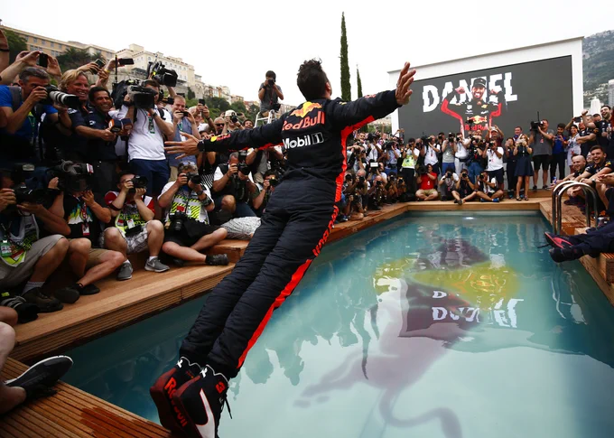 Daniel Ricciardo arches his back with his arms stretched out to the side as he is mid swan dive into a Red Bull branded swimming pool. The area is completely surrounded by photographers, press and team members, laughing as he falls in. 2018 Monaco Grand Prix.