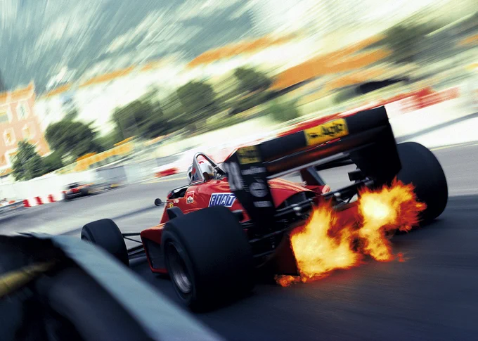 Shot using the pull zoom technique that blurs the surroundings to give a sense of speed, a rear facing view of Stefan Johansson in his red and black Ferrari 156/85, close to crash barriers on the left. Exhaust flames spit up through the rear diffuser. 1985 Monaco Grand Prix.