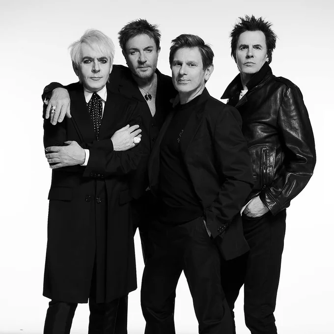 Four individuals are posing together in a black and white image. They are wearing dark, stylish clothing. The background is plain, enhancing the focus on the group.
