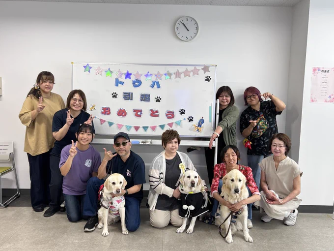室内、背景にあるホワイトボードにトウハ、引退、おめでとう！と紙で作られた貼られている。ホワイトボード前には地元のボランティアさんたち、ユーザーさん3組と盲導犬たちが並んでの記念写真。みんな笑顔で素敵な温かい風景。