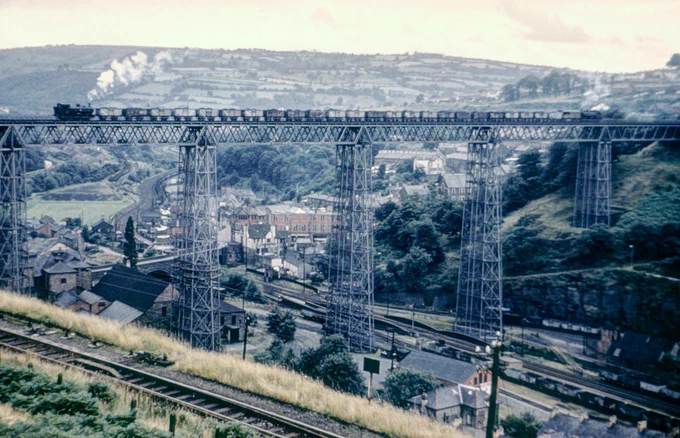 A long train of 16 ton mineral wagons with a tank engine at each end crosses the wrought iron Crumlin Viaduct, which towers above the Ebbw valley below. The vantage point is the village of Swffryd, the still surviving line to Ebbw Vale can be seen in the valley bottom while the connecting line between the high and low level routes can be seen in the foreground.