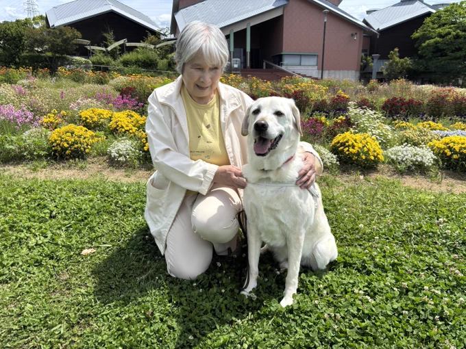 公園でお花畑を拝見に盲導犬とユーザーさんが座っての記念写真。緑とピンクや黄色のお花が綺麗。盲導犬もユーザーんも笑顔。