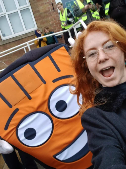 Person with curly red hair takes a selfie next to a large, smiling orange cartoon character costume outdoors near a brick building.
