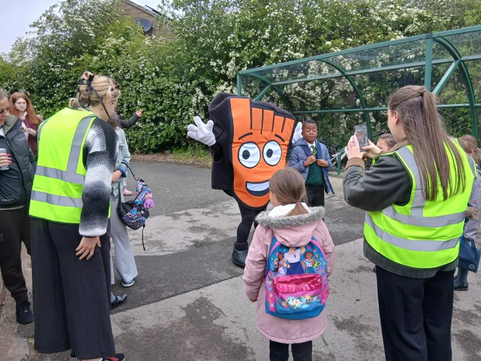 A person in a large orange mascot costume waving to children near a school bus shelter while adults in safety vests supervise.