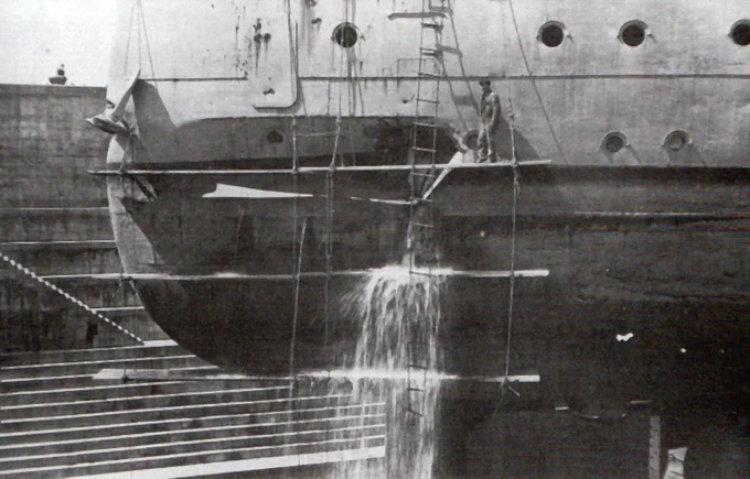 With HMS Warspite in dry dock at Rosyth after the Battle of Jutland, a dockyard worker stands beside the hole in the battleship's stern, signifying the shell hit that damage her steering during a hectic fight with the enemy battle fleet. Image: Franklin Collection, as used in 'Warspite' by Iain Ballantyne.