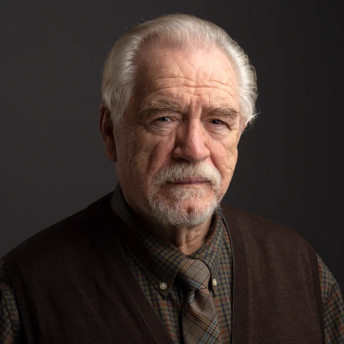 Actor Brian Cox looks intently into the camera. He wears a dark brown waistcoat with a checked shirt and tie.