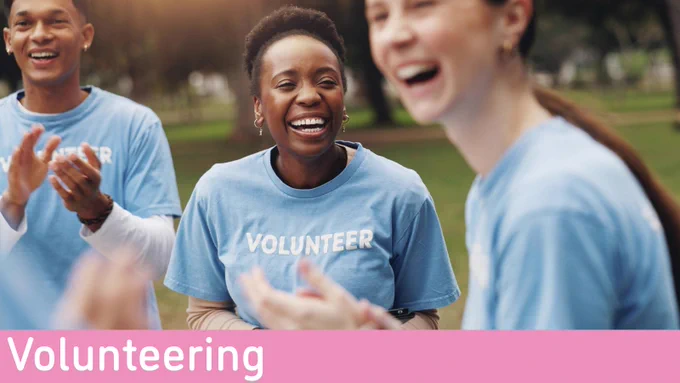 Three people wearing blue "VOLUNTEER" shirts are clapping or talking outdoors. A pink banner in the bottom reads "Volunteering"