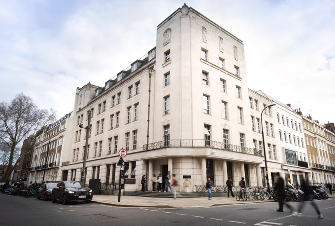 The street view of Bentham House, home to UCL Faculty of Laws. Bentham House is a large, white stone building on a corner, featuring multiple windows and a classic design. People and bicycles are in the foreground.