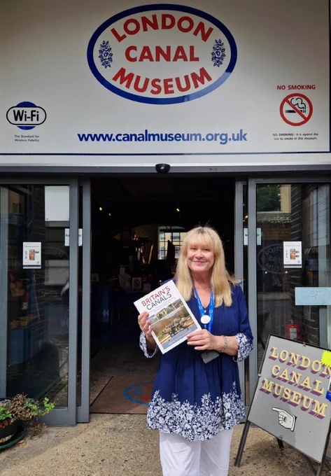 A museum volunteer stands outside the London Canal Museum holding a copy of Britain’s Canals by Anthony Burton and Derek Pratt, June’s book recommendation. The museum entrance and signage are visible in the background.