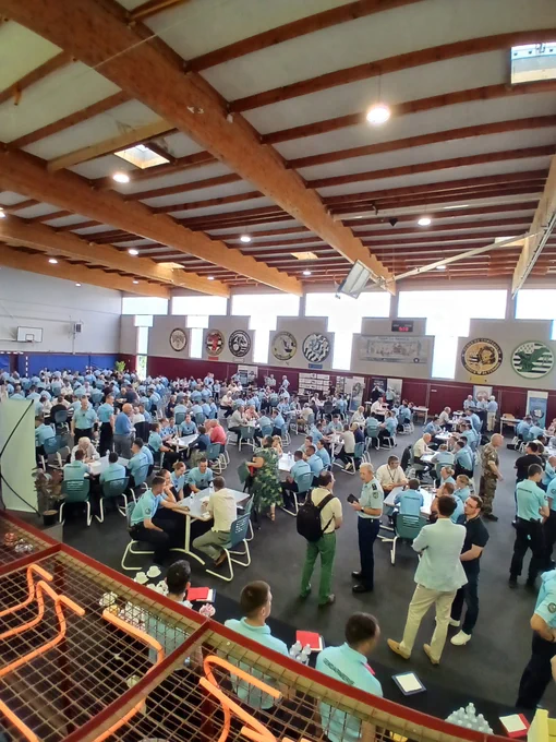 Vue d'ensemble d'un grand gymnase rempli de nombreuses personnes portant pour la plupart des polos bleu clair, assises à des tables rondes ou debout en train de discuter. L'ambiance est celle d'une réunion professionnelle ou d'un forum, avec beaucoup d'activité et d'échanges. Les murs sont décorés d'insignes et de logos, et la lumière naturelle entre par de grandes fenêtres en hauteur.