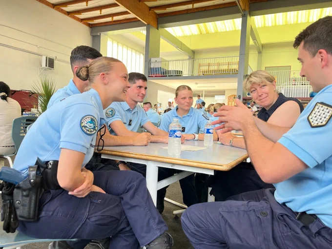 Un groupe de six personnes, dont cinq en uniforme bleu clair de gendarmerie et une femme en civil, sont assis autour d'une table dans une grande salle lumineuse. Trois bouteilles d'eau sont posées sur la table. Les membres du groupe discutent et sourient, semblant partager un moment convivial. L'arrière-plan montre d'autres personnes assises et une mezzanine sous un toit vitré.